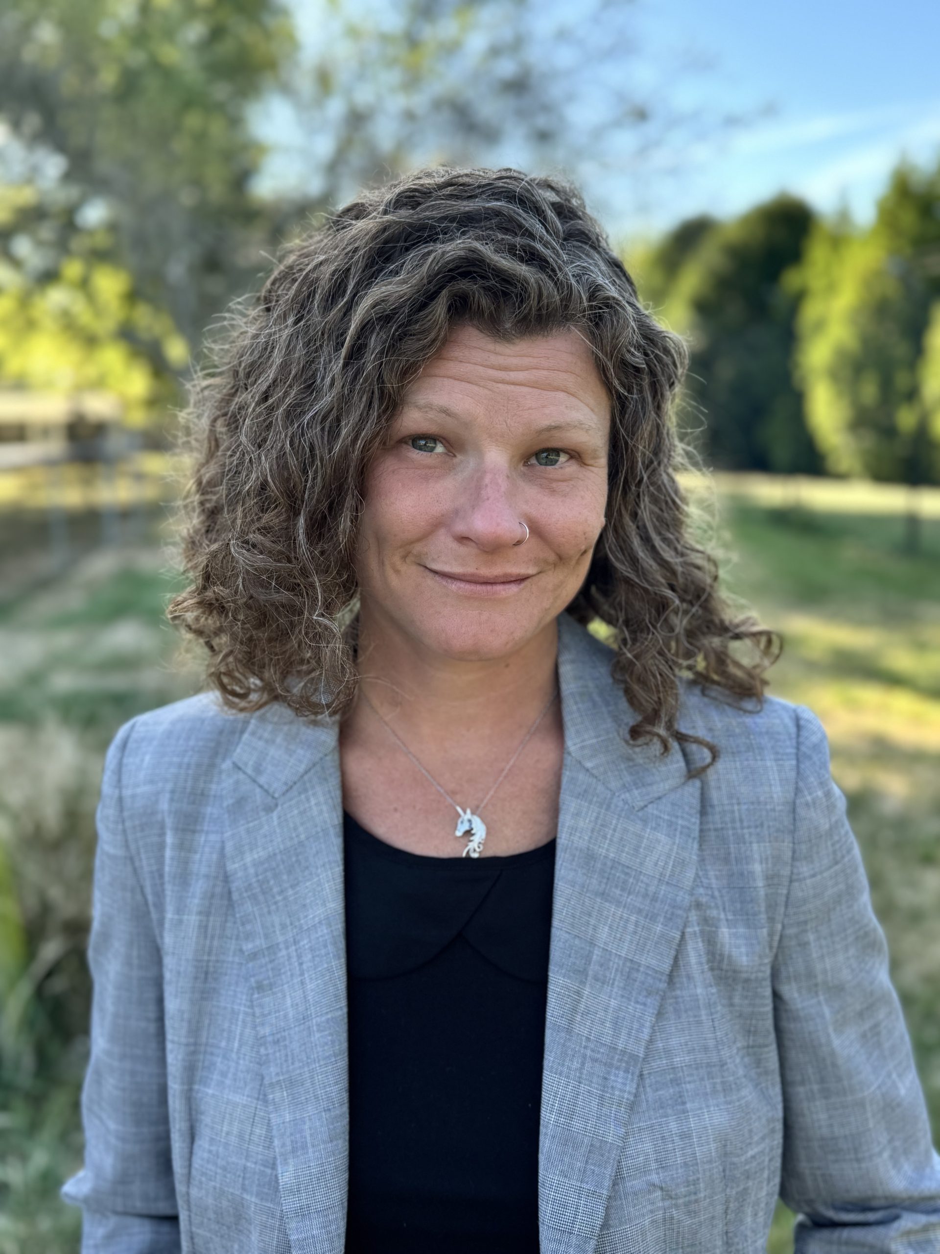 white woman with brown curly hair photographed from the chest up wearing a grey blazer and black shirt and smiling outside
